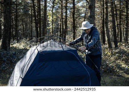 A lone explorer is seen installing his tent amidst the tranquil forest setting as the sun sets, ready for a delightful summer night spent immersed in nature's embrace