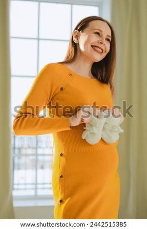 Pregnancy motherhood people expectation future. Pregnant woman with big belly holding newborn baby booties smiling at home. Young mom enjoying pregnancy. Maternity tenderness parenthood new life Royalty-Free Stock Photo #2442515385