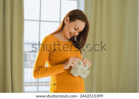 Pregnancy motherhood people expectation future. Pregnant woman with big belly holding newborn baby booties smiling at home. Young mom enjoying pregnancy. Maternity tenderness parenthood new life Royalty-Free Stock Photo #2442515399