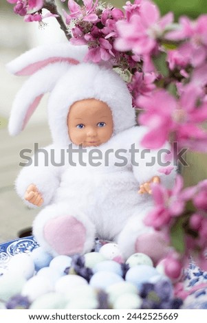 Baby doll in a white Easter bunny costume, against a background of sakura flowers and Easter eggs, Easter concept, High quality photo