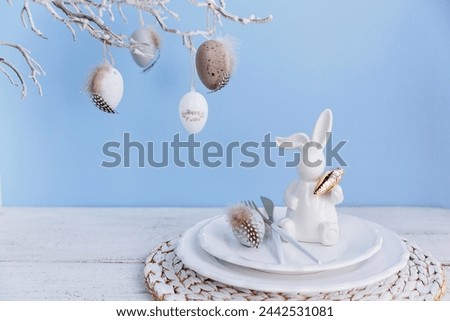 Table place setting with white bunny figure and eggs and festive easter decor on pastel blue background. Happy easter greeting or invitation card