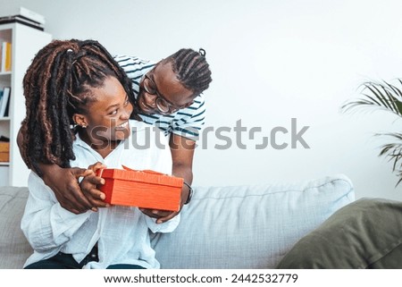 Surprised woman receiving Birthday present from her boyfriend. Young happy woman being surprised by her boyfriend with a birthday preset. Happy couple with gift box hugging at home.
