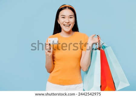 Young fun woman of Asian ethnicity wears orange t-shirt casual clothes hold package bags after shopping hold credit bank card isolated on plain blue cyan background. Black Friday sale buy day concept