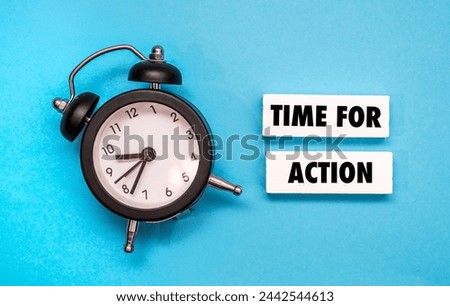 A black alarm clock sitting next to coloured wooden block with word “TIME FOR ACTION” on it