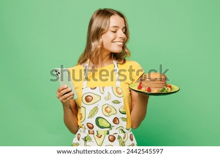 Young cool housewife housekeeper chef cook baker woman wear apron yellow t-shirt hold in hand plate with pancakes use mobile cell phone isolated on plain pastel green background. Cooking food concept