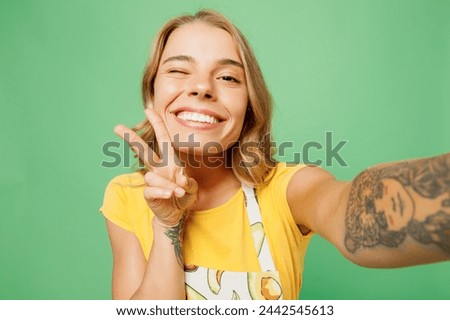 Close up young housewife housekeeper chef cook baker woman wears apron yellow t-shirt do selfie shot pov on mobile cell phone show v-sign wink isolated on plain green background. Cooking food concept