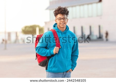 A confident student brazilian guy with a red backpack walking in an urban setting, looking at the camera Royalty-Free Stock Photo #2442548105