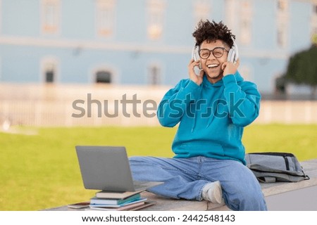 A content student brazilian guy sits with a laptop while wearing headphones, books laid out around on a sunny day outdoors Royalty-Free Stock Photo #2442548143