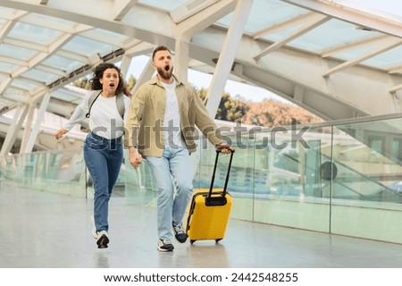 Two people in casual attire are running with expressions of panic, possibly late for a flight, in an architecturally modern airport with a yellow suitcase Royalty-Free Stock Photo #2442548255