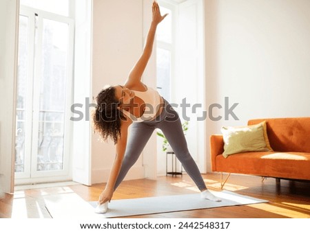 Sporty young woman in sportswear doing rotating toe touches, training flexibility and body strength at home setting. Concept of domestic fitness workout, active lifestyle and weight loss Royalty-Free Stock Photo #2442548317