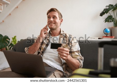 Portrait of smiling guy, tourist booking hotel, confirming purchase over the phone, holding credit card and laptop. Royalty-Free Stock Photo #2442561301
