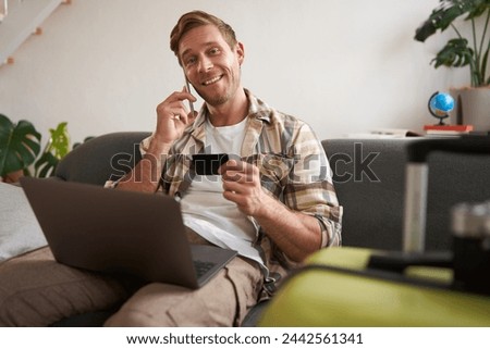 Portrait of young man with credit card, sitting and using laptop, making a phone call, booking hotel or tickets, confirming his purchase over telephone, has suitcase near him. Royalty-Free Stock Photo #2442561341