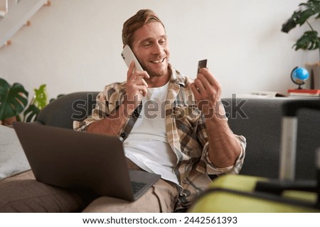 Portrait of smiling guy, tourist booking hotel, confirming purchase over the phone, holding credit card and laptop. Royalty-Free Stock Photo #2442561393