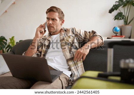 Portrait of young man with credit card, sitting and using laptop, making a phone call, booking hotel or tickets, confirming his purchase over telephone, has suitcase near him. Royalty-Free Stock Photo #2442561405