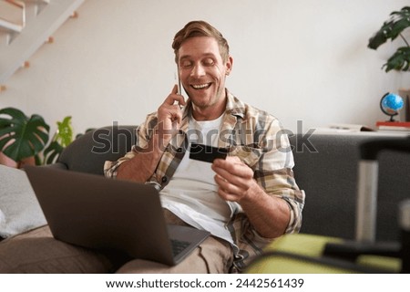 Portrait of young man with credit card, sitting and using laptop, making a phone call, booking hotel or tickets, confirming his purchase over telephone, has suitcase near him. Royalty-Free Stock Photo #2442561439