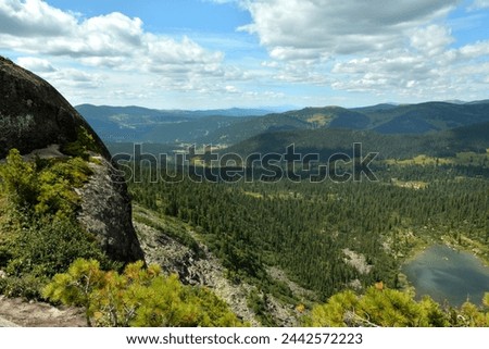 A rocky massif overgrown with grass on top of a mountain overlooking a picturesque lake in dense mountain taiga under a cloudy summer sky. Natural park Ergaki, Krasnoyarsk region, Siberia, Russia. Royalty-Free Stock Photo #2442572223
