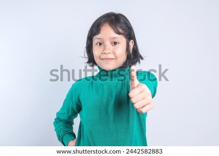 Adorable Asian little girl in green sweater showing thumb up over grey background