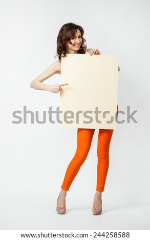 Playful young woman in orange pants holding blank placard showing at it, full length portrait on neutral background