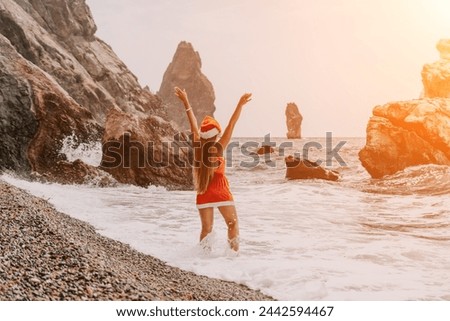Woman summer travel sea. Happy tourist in red bikini and Santas hat enjoy taking picture outdoors for memories. Woman traveler posing on the beach surrounded by volcanic mountains, sharing travel joy