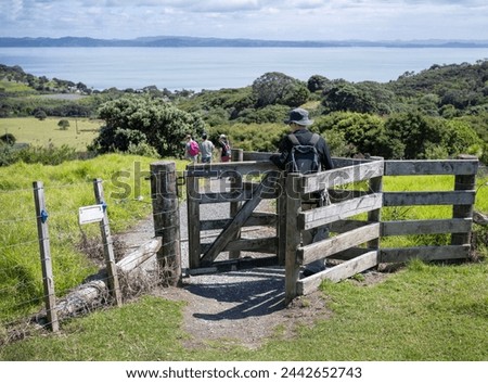 People walking through the farm gate on a hiking track. Electric  Fence sign hanging on the fence. Shakespear Regional Park. Auckland.