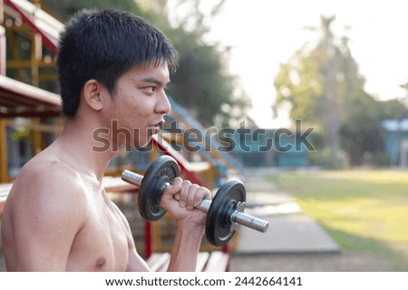 Asian teenboy holding dumbbell in left hand and lifting to do the exercise in the evening beside green grass lawn of his community, soft focus, lifestyle of teenagers concept.