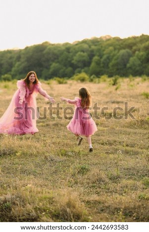 Mother and daughter girl in same designed long pink dresses posing on nature in summer. Happy daughter in beautiful designed dress running to her mother on the meadow. Love. Family concept
