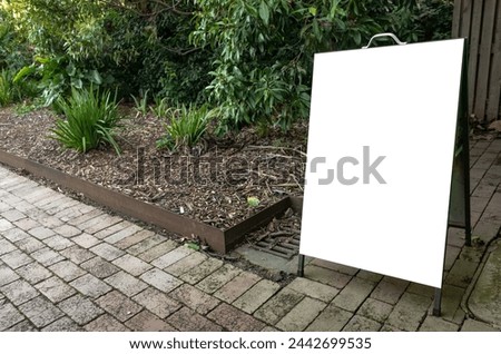 A sidewalk sign with a blank white board stands against a backdrop of lush garden greenery and a brick pavement. Empty blank Mockup background texture in natural outdoor setting.