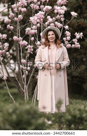 Woman magnolia flowers, surrounded by blossoming trees., hair down, white hat, wearing a light coat. Captured during spring, showcasing natural beauty and seasonal change.
