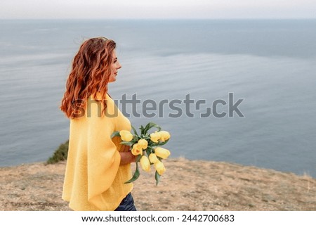 Rear view of a woman with long hair against a background of mountains and sea. Holding a bouquet of yellow tulips in her hands, wearing a yellow sweater