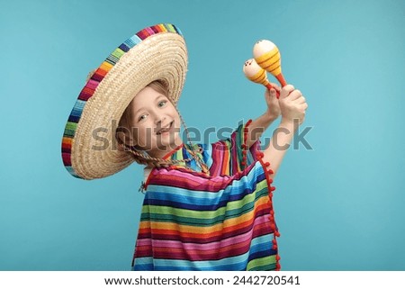 Cute girl in Mexican sombrero hat and poncho dancing with maracas on light blue background