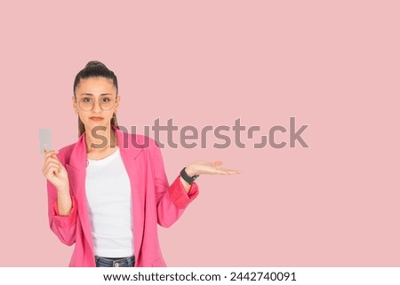 Sad disappointed young brunette 20s woman holding credit card she stretched her open palm aside copy space. Don't like this invisible product. Wearing pink jacket  standing over pastel pink background