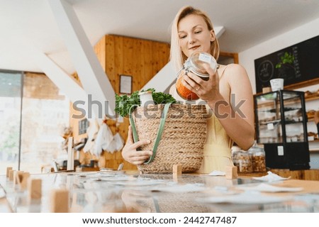 Smiling woman with glass jar of red lentils in zero waste shop. Female customer with basket of organic products in modern plastic free store. Concept of retail of eco food and sustainable shopping. Royalty-Free Stock Photo #2442747487