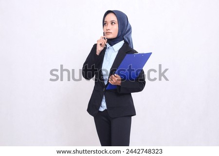 Asian mature office woman in hijab standing thinking to the right carrying a pen and hugging a clipboard. Beautiful Muslim women wearing blue shirts for business, finance and corporate use