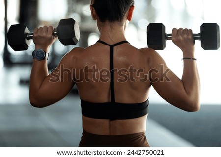 Back side, strong muscular young woman lifting dumbbell weights at the gym, doing exercises with dumbbell, fitness muscular body