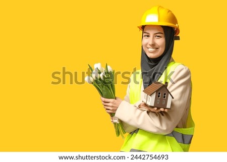 Muslim female construction worker with toy house and white tulips on yellow background. Women's Day celebration