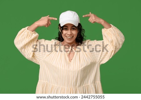 Pretty African-American woman in dress and baseball cap on green background