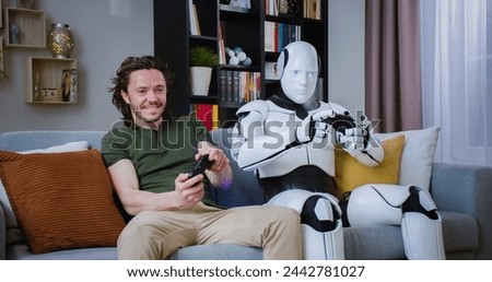 Handsome caucasian young man sitting on cozy couch with smart futuristic robot and playing video games on console. Unshaved man getting upset about losing match. Royalty-Free Stock Photo #2442781027