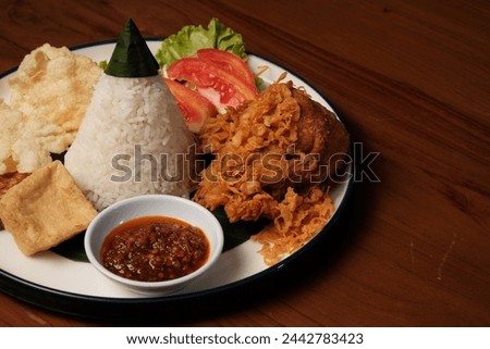 Nasi Tumpeng Ayam Goreng is a traditional Indonesian dish that presents a delicious combination of flavors and textures. The heart of this dish is the cone-shaped rice, known as "nasi tumpeng", Royalty-Free Stock Photo #2442783423