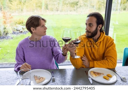 This inviting photograph captures two friends sharing a moment over wine, exchanging meaningful glances that suggest deep conversation and understanding. The casual dining experience with pizza slices Royalty-Free Stock Photo #2442795405