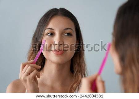 Facial hair removal. Dermaplaning. Close-up of beautiful teenager girl shaving her face by razor at home. Pretty young woman using razor on bathroom. Royalty-Free Stock Photo #2442804581