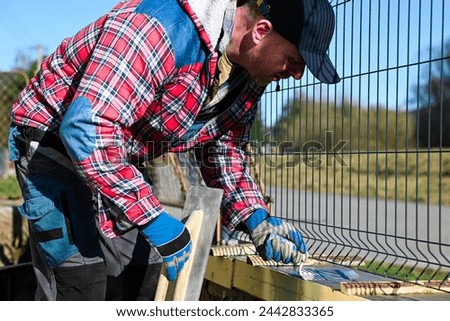 A man in overalls and gloves is repairing the fence in front of the family house. Close-up view and blurred background.