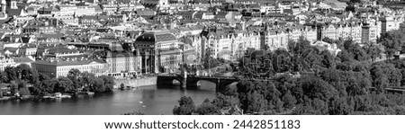 A view overlooking the National Theatre in Prague, with the Vltava River and a bridge in the foreground surrounded by trees and architecture. Prague, Czechia. Black and white photography.