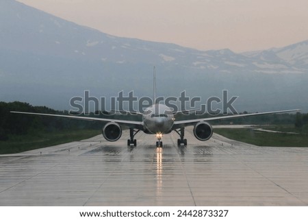Frontal view of Aircraft coming in for Landing against background of Snow-capped Mountain Peaks on Rainy cloudy day. The Light Path from Headlights creeps along the Wet runway towards the airport. Royalty-Free Stock Photo #2442873327
