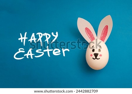 Food photo for Easter holiday. Chicken egg with cute rabbit face and bunny ears on blue background. Greeting card. Happy Easter handwritten white text. Preparation for celebration. Family holiday