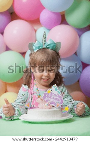 A little girl in a unicorn dress and hairband is holding a pink unicorn soft toy and looking an her birthday cake. Four years-old celebration. Pastel latex balloons background. Vertical image. 