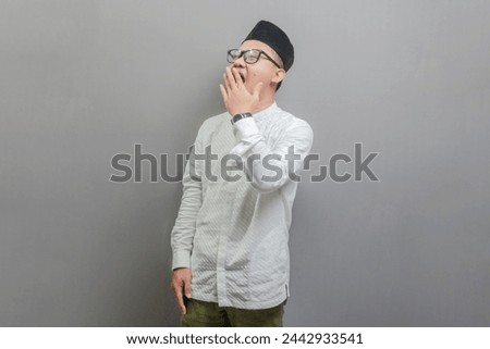 Asian Muslim man wearing a koko shirt and peci with shades of the fasting month, yawning and covering wide open mouth with hand, isolated on a gray background
