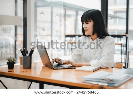 Smiling businesswoman multitasks, using a smartphone and laptop at a spacious cafe table, surrounded by notes and reports.