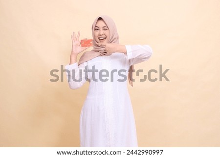 Asian Muslim woman wearing a hijab smiling cheerfully, winking, holding a debit credit card in her hand and pointing up. Lifestyle, business and finance concept