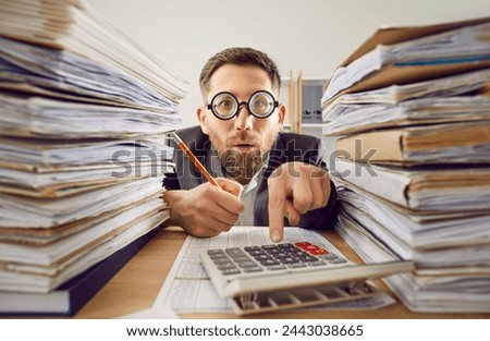 Overworked business man in suit and funny glasses working at the desk on his workplace at office with a pile of folders and a stack of papers. Tired accountant making calculations analyzing data. Royalty-Free Stock Photo #2443038665