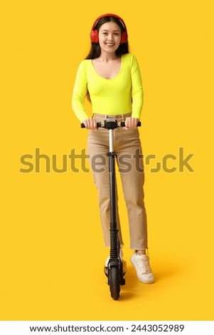 Beautiful young Asian woman in headphones with modern electric kick scooter on yellow background